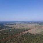 Aerial view of a vast rural landscape with fields, trees, and a small pond. A red outline marks the boundaries of a large plot of land under a clear blue sky.