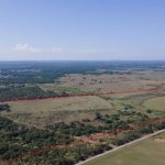 Aerial view of a large, open rural landscape with fields, trees, and scattered vegetation. A red outline marks the boundary of a specific parcel of land. A road runs along one side of the outlined property.