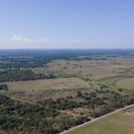 Aerial view of a vast, open rural landscape with green fields, scattered trees, a small pond, and a road running along the bottom right; a clear blue sky stretches above the horizon.