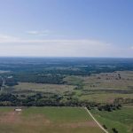 Aerial view of a vast rural landscape with green fields, patches of trees, small ponds, dirt roads, and a clear blue sky stretching across the horizon.