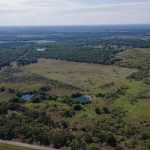 Aerial view of a vast rural landscape with open green fields, scattered trees, small ponds, and a road running along the bottom edge under a partly cloudy sky.