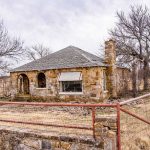 A small, abandoned stone house with a shingled roof stands behind a rusted wire fence on the Historic S&S Ranch, nestled in a dry, grassy field and surrounded by leafless trees under a cloudy sky.
