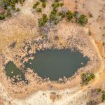 Aerial view of a small, irregularly shaped pond at Historic S&S Ranch, surrounded by dry, brown grass and sparse green shrubs in a mostly arid landscape. Dirt paths wind around the pond.