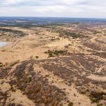 Aerial view of the historic S&S Ranch reveals a dry, hilly landscape with sparse vegetation, meandering dirt roads, patches of bushes, and a small pond on the left side beneath a cloudy sky.