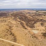 Aerial view of the Historic S&S Ranch reveals a vast, dry, rolling landscape with patches of brown grass, scattered shrubs, small ponds, and a dirt road winding through the terrain under a cloudy sky.