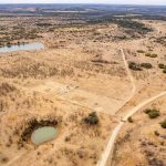 Aerial view of the dry, sparsely vegetated landscape of the historic S&S Ranch, with dirt roads, small ponds, and scattered bushes beneath a hazy sky. The terrain is mostly flat with gentle hills in the distance.