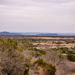A wide landscape of dry, grassy plains with scattered bushes and trees under a cloudy sky stretches across the S&S Ranch, with distant low hills visible on the horizon.