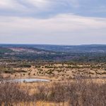 A wide, open landscape under a cloudy sky at the historic S&S Ranch, featuring rolling hills with scattered shrubs and bushes, a small pond, and dry grasses in the foreground.