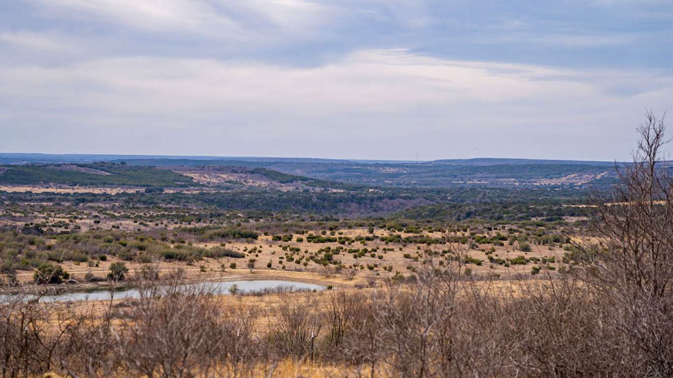 A wide, open landscape under a cloudy sky at the historic S&S Ranch, featuring rolling hills with scattered shrubs and bushes, a small pond, and dry grasses in the foreground.