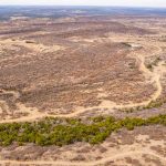 Aerial view of the historic S&S Ranch reveals a dry, expansive landscape with winding dirt roads, scattered trees, brown patches of grass, and a small pond in the lower left corner under a hazy sky.