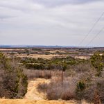 A dirt path winds through dry, bushy terrain on Historic S&S Ranch, with scattered trees under a cloudy sky. Power lines cut diagonally across the landscape, and distant hills rise on the horizon.