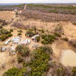 Aerial view of the Historic S&S Ranch, featuring several RVs, small buildings, scattered trees, dirt roads, and a small pond—all surrounded by open fields and patches of brush in a dry landscape.