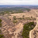Aerial view of a dry, semi-arid landscape with sparse vegetation, patches of green trees, and a winding dirt road leading through the terrain under a cloudy sky at S&S Ranch 1.