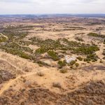 Aerial view of a vast, dry landscape with scattered green shrubs and trees, winding dirt paths, and the historic S&S Ranch nestled near the center right. The distant horizon stretches beneath a cloudy sky.
