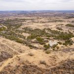 Aerial view of the vast, dry landscape surrounding the historic S&S Ranch, with green shrubs and trees, dirt paths weaving through the terrain, and a small cluster of buildings near the center-right under an overcast sky.