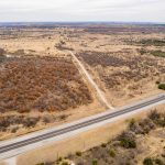 Aerial view of a rural landscape with a paved road in the foreground and a dirt road leading into dry, scrubby fields surrounding the historic S&S Ranch, with rolling hills stretching under a cloudy sky.