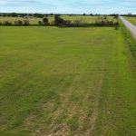 A wide, open grassy field stretches alongside a straight rural road, with trees and scattered buildings visible in the distance under a mostly cloudy sky.