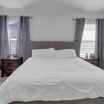 A neatly made bed with white bedding is centered between two windows with gray curtains. Nightstands flank the bed, a white armchair sits on the left, and a white baby crib is on the right. The walls are light gray.