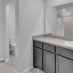 Modern bathroom with a large mirror, single sink, gray cabinets, and granite countertop. To the left, an open door leads to a walk-in closet with clothes, shoes, and shelves. A toilet and shower are also visible.