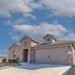 A modern, single-story brick house with a two-car garage and dormer windows, set on a neatly paved driveway, with a clear blue sky and a few clouds overhead.