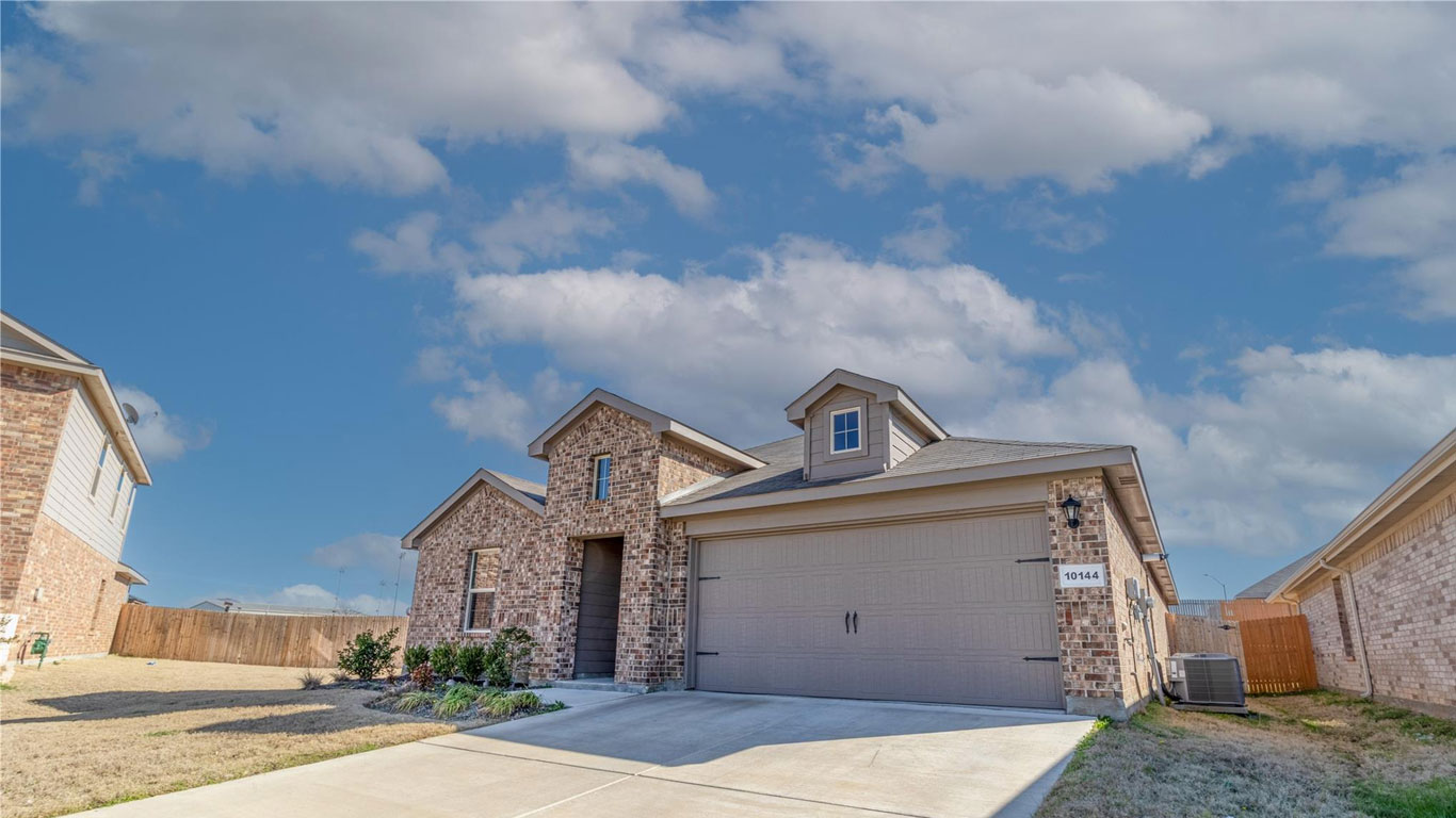 A modern, single-story brick house with a two-car garage and dormer windows, set on a neatly paved driveway, with a clear blue sky and a few clouds overhead.