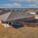 Aerial view of a suburban backyard with a fenced yard, covered patio, and grill. Surrounding houses with gray rooftops are visible, along with a street and several parked cars in the neighborhood.