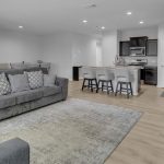 Modern open-concept living space with gray sofas, a patterned rug, a large TV, and a kitchen with a breakfast bar and upholstered stools. Light wood flooring and neutral walls throughout. Dining area visible in the background.