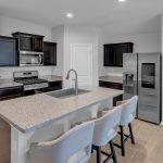 Modern kitchen with dark cabinets, stainless steel appliances, a center island with a sink, and three upholstered barstools. Light wood floors and a small kids' table with two chairs are in the corner.