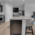 Modern kitchen with dark cabinets, stainless steel appliances, and a large island with bar stools; open layout leads to a living area with a TV on a cabinet and light wood flooring throughout.
