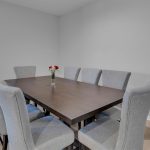 A modern dining room with a dark wooden table surrounded by eight light gray upholstered chairs. A small vase with red and white flowers sits in the center of the table. The walls are light gray and the floor is light wood.