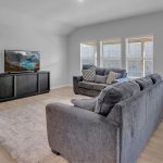 A modern living room with light wood flooring, gray sofas, a TV on a black stand, a large area rug, and three tall windows. A small white kids’ table and chairs are in the corner near the TV.