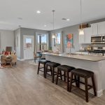 Open-concept kitchen and living room with a large island, white cabinets, stainless steel appliances, and bar stools. Adjacent dining and seating areas are decorated in neutral tones with large windows letting in natural light.