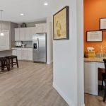 Modern kitchen with white cabinets, stainless steel appliances, and a breakfast bar with stools; a small office nook with a built-in desk, orange accent wall, lamp, and framed calendars is visible to the right.