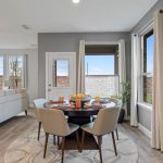 Modern dining area with a round table set for four, beige chairs, and a bowl of fruit. Large windows and a glass door let in natural light; abstract orange artwork hangs on a gray wall. A living room is visible in the background.