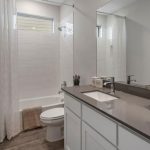 Modern bathroom with a gray countertop, white cabinets, a sink, a toilet, a bathtub with a white tile surround, a window above the tub, and decorative artwork and plants.
