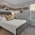 A modern bedroom with a wooden bed frame, white bedding, patterned pillows, two bedside tables with lamps, abstract wall art, and a window with striped curtains letting in natural light.