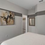 A modern bedroom with light gray walls, a bed with a chevron-patterned pillow, abstract wall art, a mirror, and a white door. The room has minimal décor and a clean, neutral color scheme.