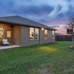 A modern single-story house with brown siding and large windows sits on a well-kept lawn. A small covered patio is visible. The sky is vibrant with sunset colors and a wooden fence encloses the backyard.