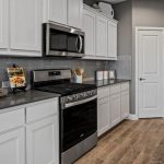 Modern kitchen with white cabinets, stainless steel appliances, gray subway tile backsplash, light wood flooring, and gray countertops. Decorative items and kitchen essentials are arranged on the counters.