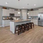Modern kitchen with light wood flooring, white cabinets, a central island with three dark stools, stainless steel appliances, and pendant lights. Dining area with art on the wall is visible to the left; entryway in the background.