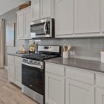 Modern kitchen with white cabinets, stainless steel gas stove and microwave, gray tile backsplash, and gray countertops. Dining area with table and chairs is visible near large windows in the background.