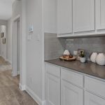 A hallway with light wood floors, white walls, and white cabinets. A gray countertop holds decorative items and baskets. A console table with a lamp and decor sits along the hallway, leading to a front door with glass panels.