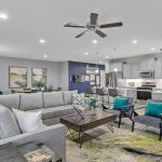 Modern open-concept living room and kitchen with gray sectional sofa, colorful pillows, two armchairs, a coffee table, and wall art. The kitchen features gray cabinets, stainless appliances, and pendant lighting.