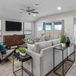 Modern living room with gray sectional sofa, accent chairs, potted plants, and a TV on the wall. Large windows let in natural light; dining area is visible in the background. Neutral tones with green accents throughout.