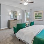 A bright bedroom with a large bed in green bedding, a green loveseat at the foot, a dresser with a mirror, two windows with curtains, a ceiling fan, and a framed bird artwork on the wall.