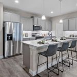 Modern kitchen with gray cabinets, stainless steel appliances, and a large island with three black barstools. The space is bright with pendant lights and has wood-style flooring. The kitchen opens to a living area.