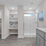 Modern bathroom with gray cabinets, a large white countertop, glass shower, open shelves with towels, and a separate toilet area; neutral colors and tile flooring throughout.
