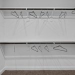 A walk-in closet with white walls, dark wooden rods on three sides, several empty black hangers, and brown carpeted flooring. The closet is clean and well-lit.