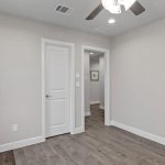 A small, empty room with light beige walls, dark wood flooring, a ceiling fan with lights, and two white doors. A window with patterned curtains is on the left side of the room.