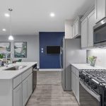 Modern kitchen with gray cabinets, stainless steel appliances, a gas stove, white countertops, and a center island. Open layout leads to a dining area with green chairs and wall art, and a blue accent wall by the refrigerator.